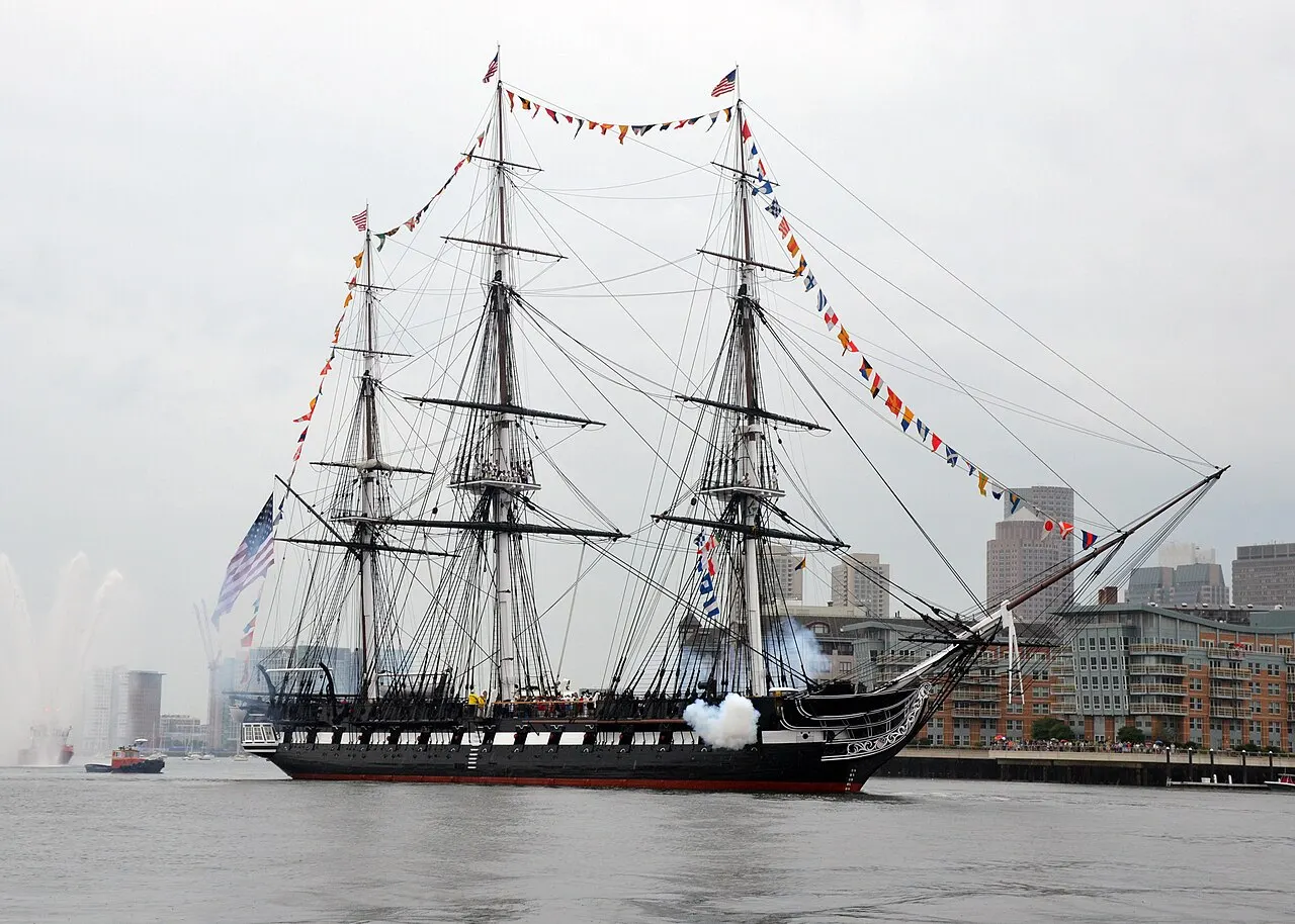 La fragata USS Constitution en el agua, con sus tres mástiles, vergas y banderas, en un día nublado.