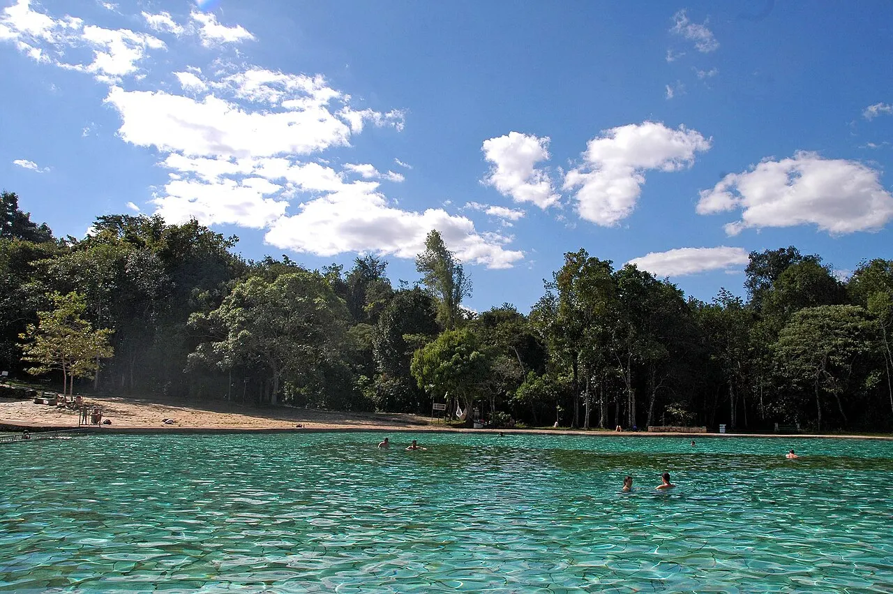 Piscina larga de água verde-clara e transparente no Parque Nacional de Brasília, com algumas pessoas nadando, faixa de areia na margem e a mata de cerrado fechada atrás, sob céu azul.