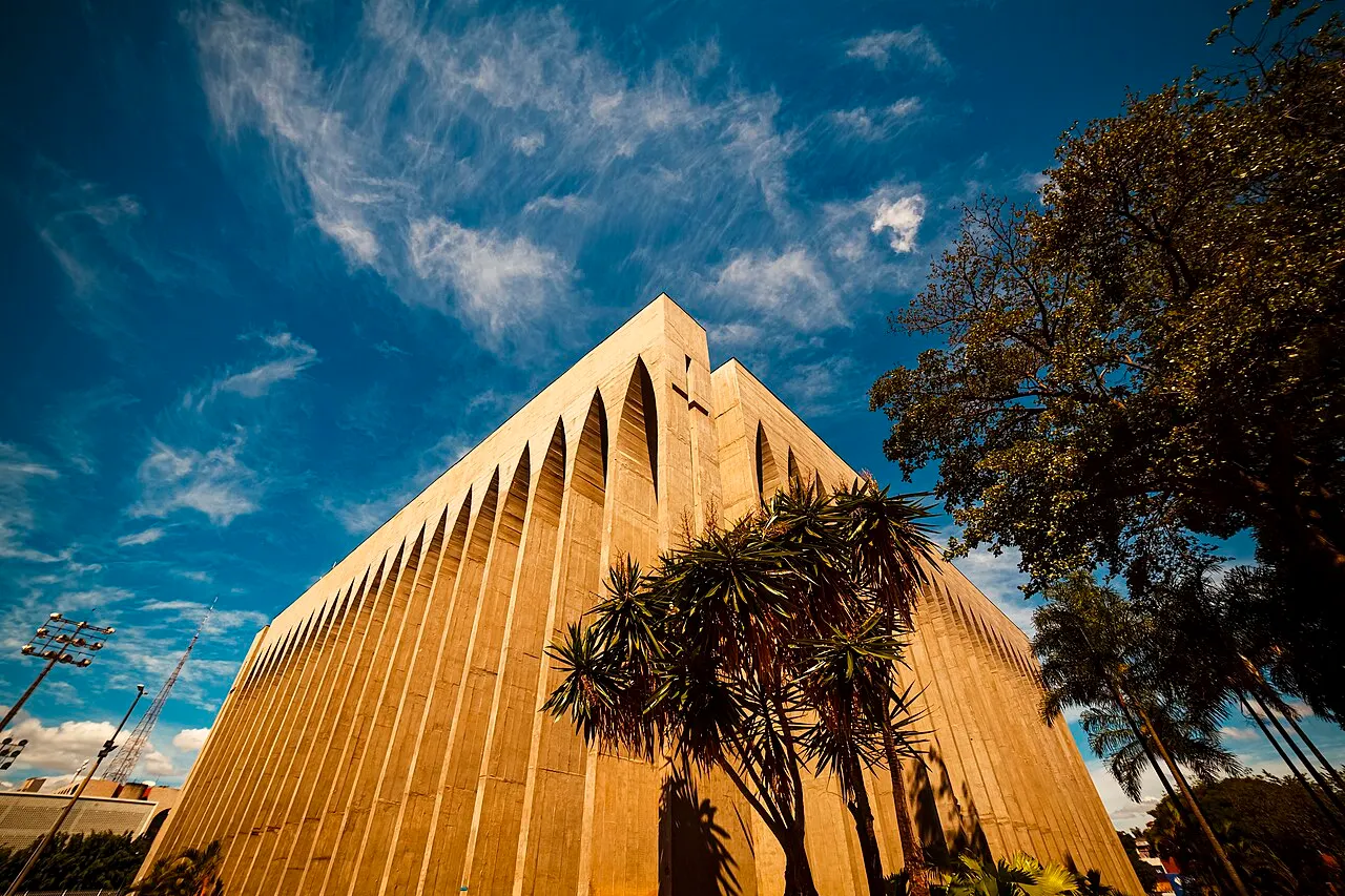 O volume de concreto do Santuário Dom Bosco sobe em frestas altas em arco, entre árvores e um céu azul.