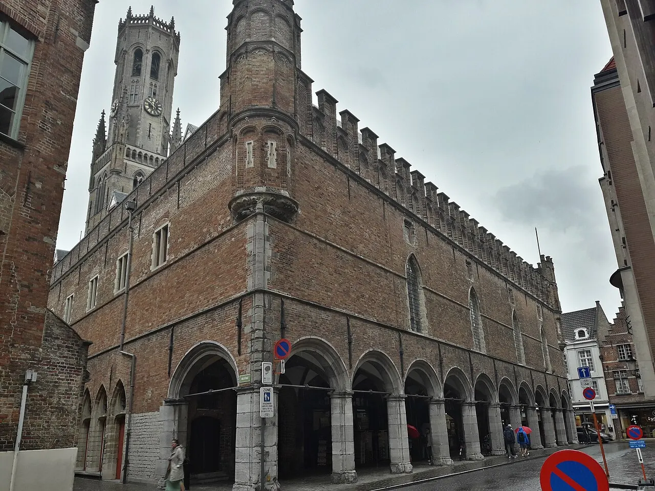O Belfort de Bruges, torre alta de tijolo e pedra, sobre os Halles com arcadas no térreo, visto da rua num dia nublado.