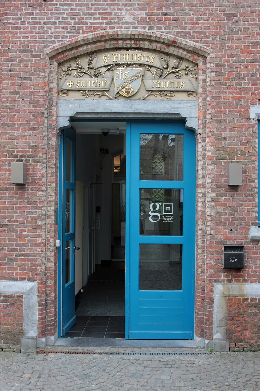 A entrada do Groeningemuseum, uma porta de vidro com moldura azul num muro de tijolo, sob uma verga de pedra entalhada.