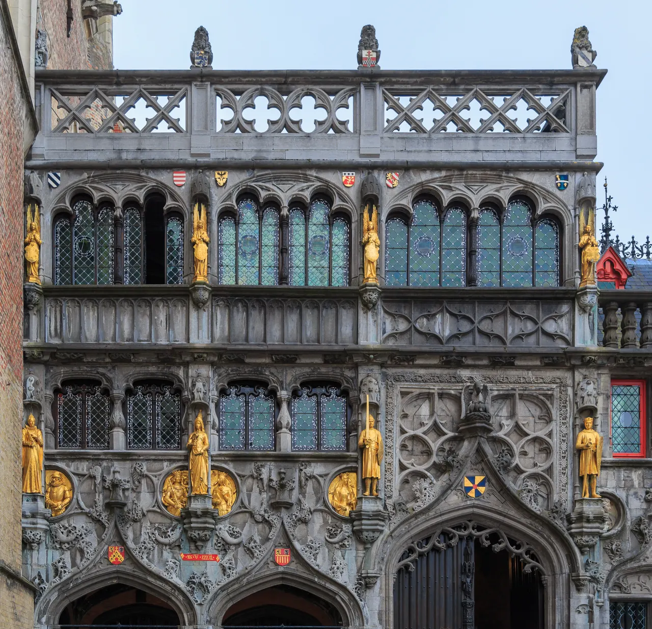 Detalhe da fachada de pedra da Basílica do Sangue Sagrado, em Bruges, com janelas de vidros coloridos, esculturas douradas, brasões e arcos ornamentados.