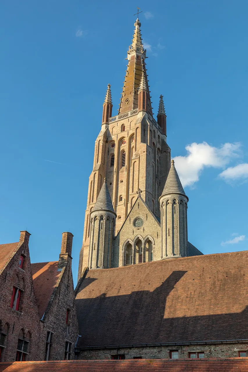 A torre alta de tijolo da Onze-Lieve-Vrouwekerk, com a ponta de pedra, acima dos telhados vermelhos, contra o céu azul.