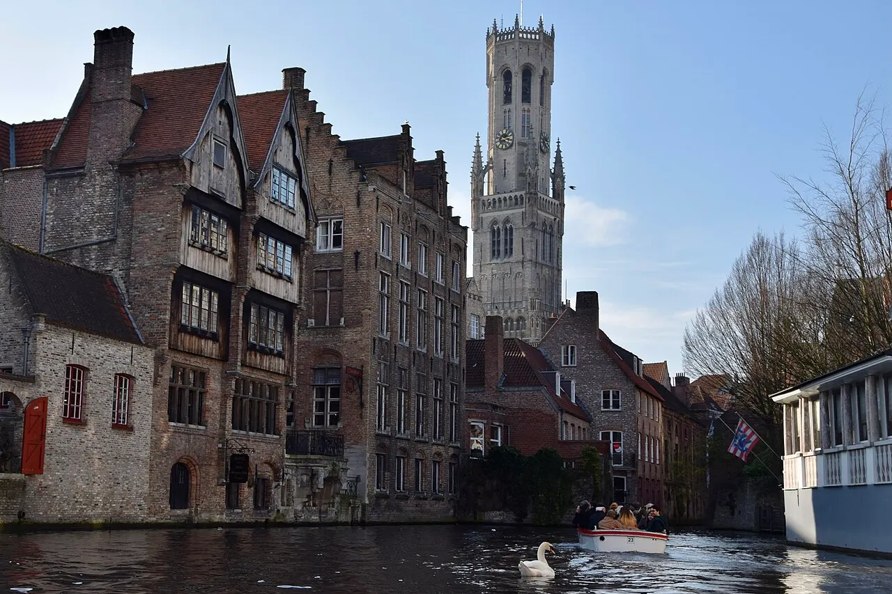 O Rozenhoedkaai: casas antigas de tijolo à beira do canal e a torre do Belfort atrás delas, com um barco de passeio à direita.