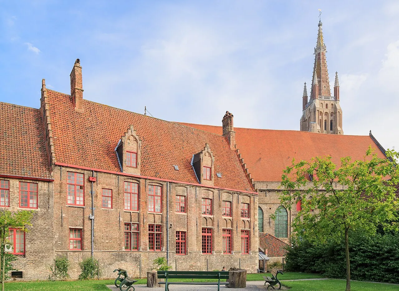 Os prédios de tijolo e telhado vermelho do antigo Sint-Janshospitaal, com um jardim à frente e a torre da Onze-Lieve-Vrouwekerk atrás.
