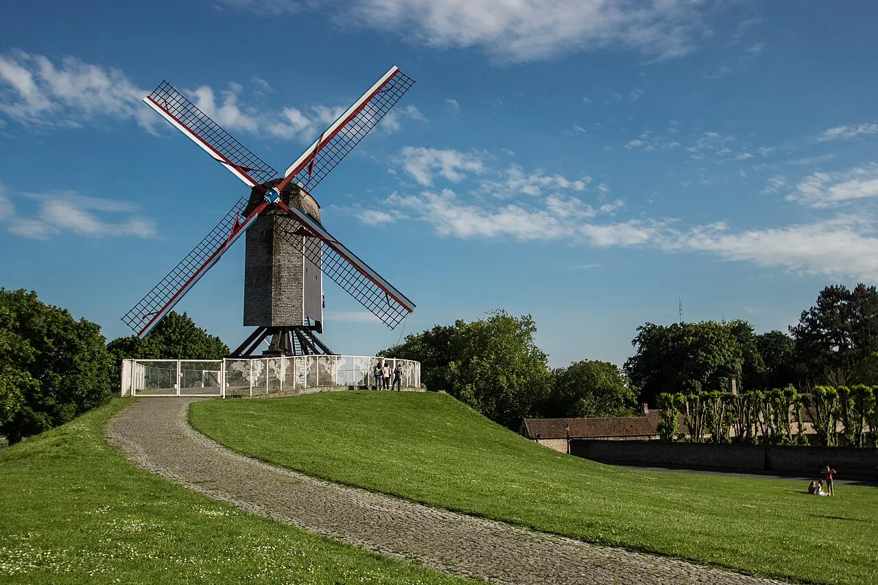 O moinho de madeira Sint-Janshuismolen no topo de um morro gramado da antiga muralha, com um caminho subindo até ele e céu azul.