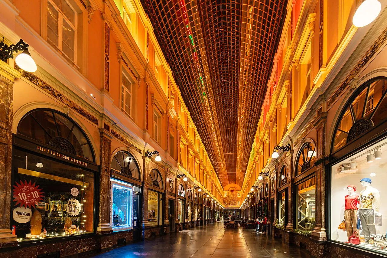 A galeria coberta das Galeries Royales Saint-Hubert segue sob a abóbada de vidro, com vitrines dos dois lados e o piso refletindo a luz.
