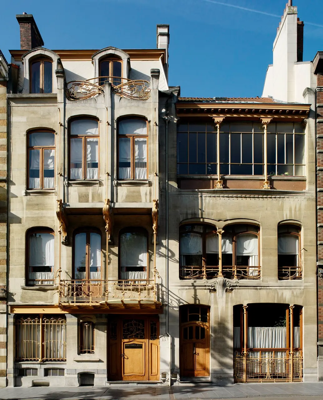 As fachadas de pedra clara de duas casas vizinhas em estilo art nouveau, com janelas de sacada, ferragens douradas curvas, portas de madeira e cortinas brancas, sob céu azul.