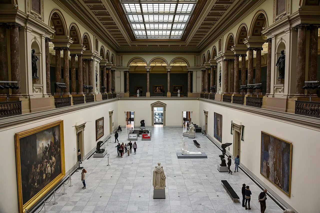 O interior de uma galeria de museu de dois andares, com colunas de mármore escuro, quadros de moldura dourada e esculturas expostas no piso térreo, claraboia de vidro no teto e visitantes circulando.