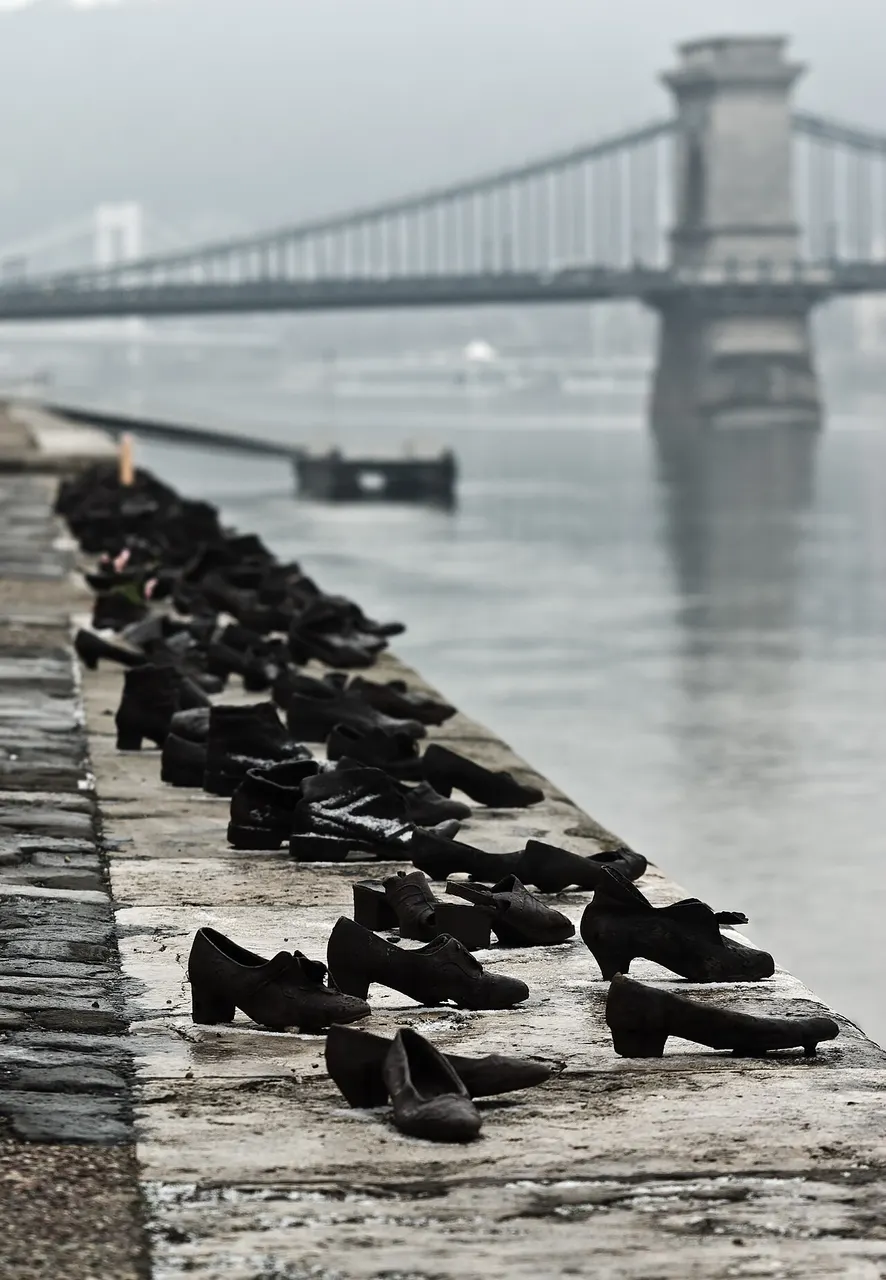 Sapatos de metal enfileirados na margem do Danúbio, em Budapeste, com uma ponte na névoa ao fundo.