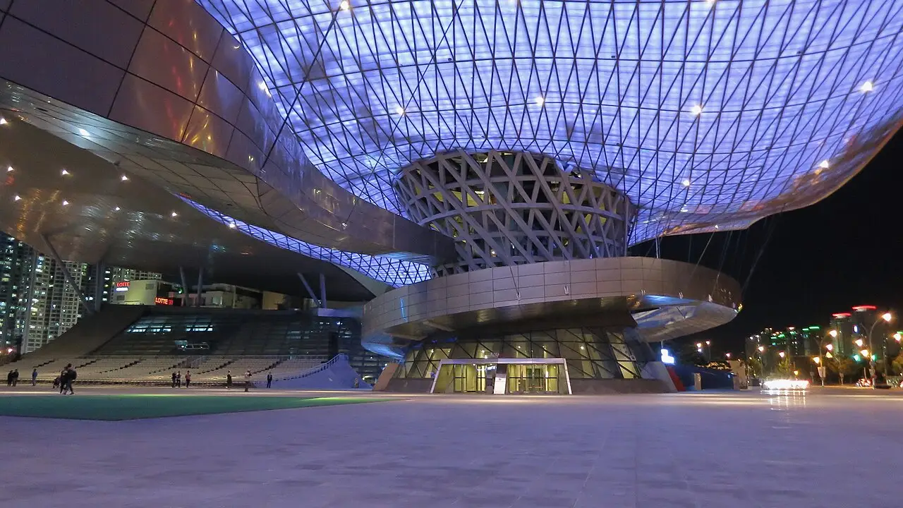 Busan Cinema Center à noite, com a cobertura de treliça azul sobre o volume curvo e a praça na frente.