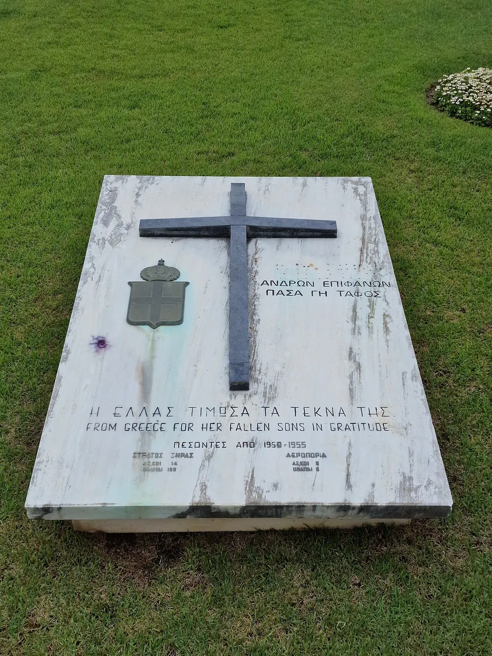 Marco de pedra branca em forma de cruz, sobre gramado verde, com placa aos pés.