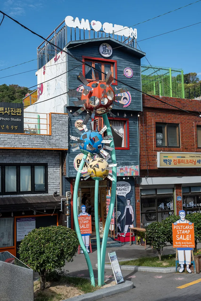 Escultura vermelha alta com cabeça mascarada colorida e pernas finas de metal, junto a uma casa da vila.