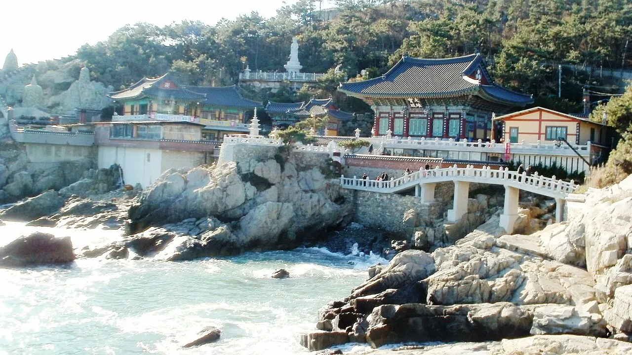 Pavilhão de telhado azul sobre rochas à beira-mar, ligado por uma ponte de pedra com estátuas de leão.
