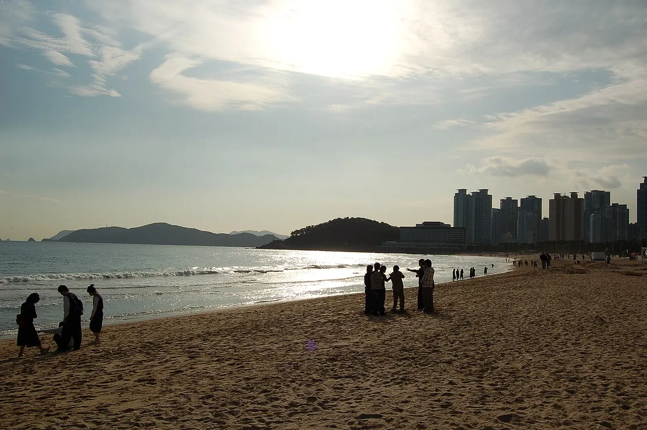 Faixa de areia em Haeundae, com pessoas em silhueta, o mar brilhando e torres na orla.