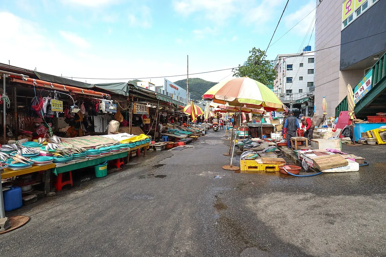 Uma rua de mercado de peixes ao ar livre, com bancas cobertas de peixe fresco sobre bandejas, baldes e caixas plásticas, guarda-sóis coloridos abertos e colinas ao fundo, sob céu parcialmente nublado.