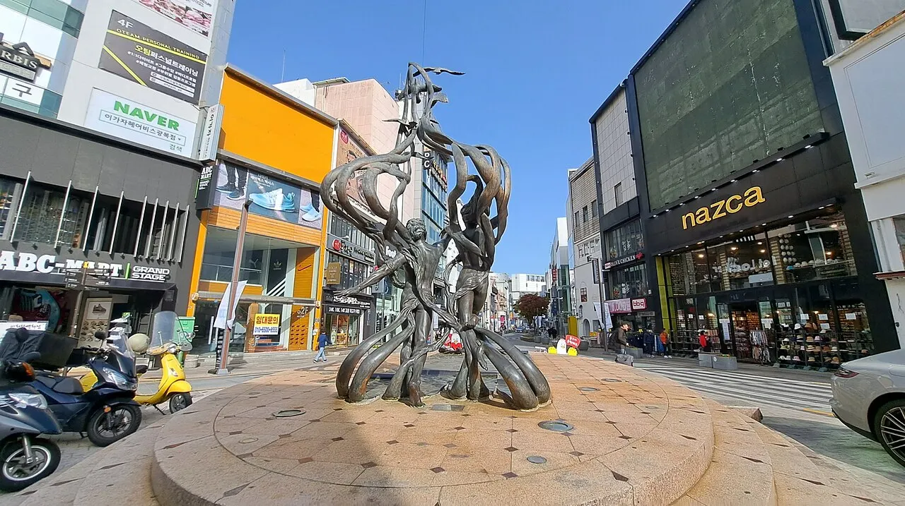 Escultura metálica alta e sinuosa, com um pequeno estandarte no topo, numa praça cercada de lojas.