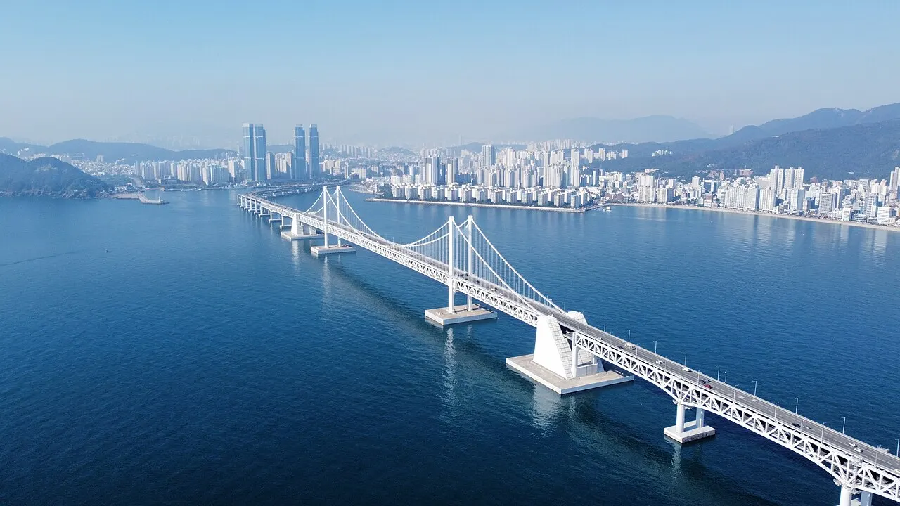 Vista aérea da ponte suspensa Gwangan em dois tabuleiros sobre a água azul da baía de Busan, com a praia e uma faixa de arranha-céus na margem ao fundo.