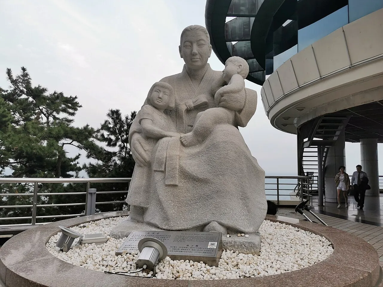 Estátua de pedra de uma mulher sentada com uma criança no colo, num mirante rochoso à beira-mar.