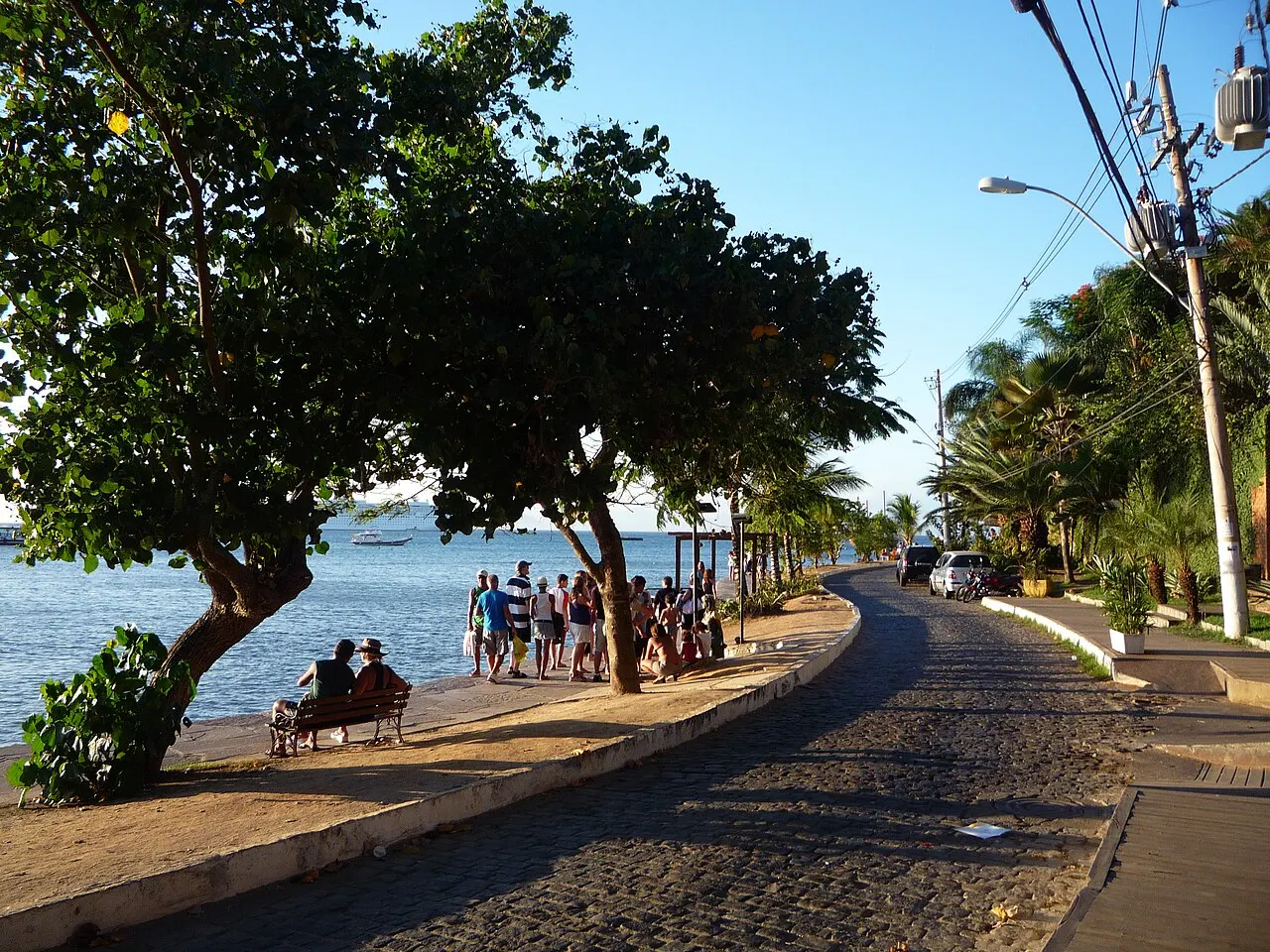 O calçadão de pedra da Orla Bardot acompanhando a beira da água, com árvores dando sombra e gente sentada no muro.