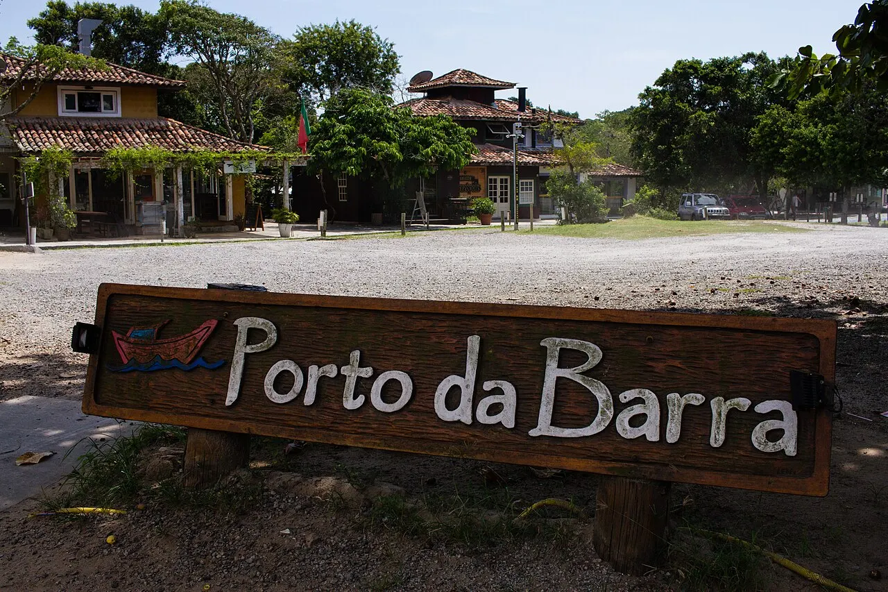 A placa de madeira do Porto da Barra na areia, com os restaurantes entre as árvores ao fundo.