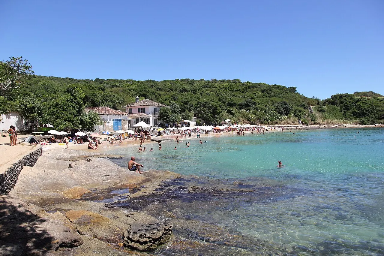 A água rasa e clara da Praia Azeda sobre as pedras, com a faixa de areia e a mata do morro ao fundo.
