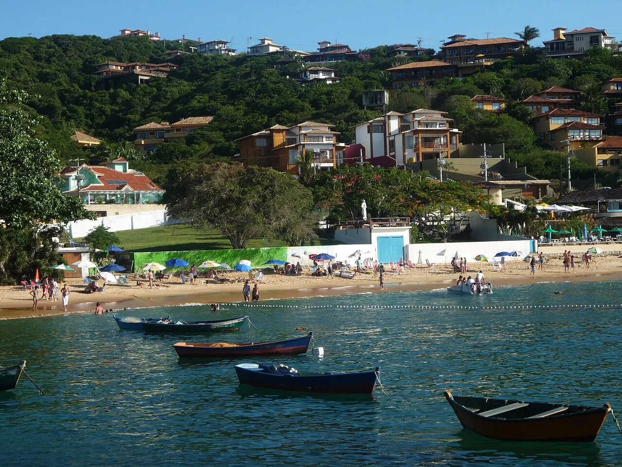 Small boats on the green water of the João Fernandes cove, with the beach and hillside houses behind.