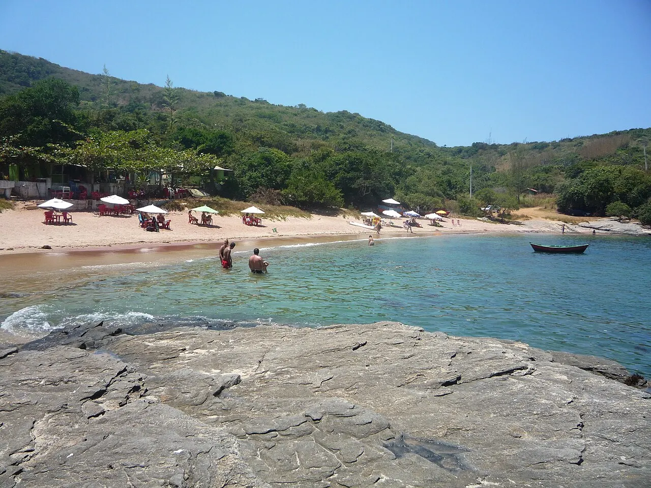 A laje de pedra cinza na frente e a enseada da Praia do Forno, com guarda-sóis na areia e morros verdes.