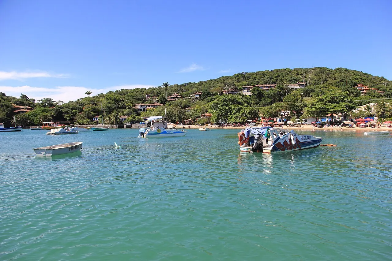 Boats moored in the green water of the Praia dos Ossos cove, with the hill of houses and trees behind.