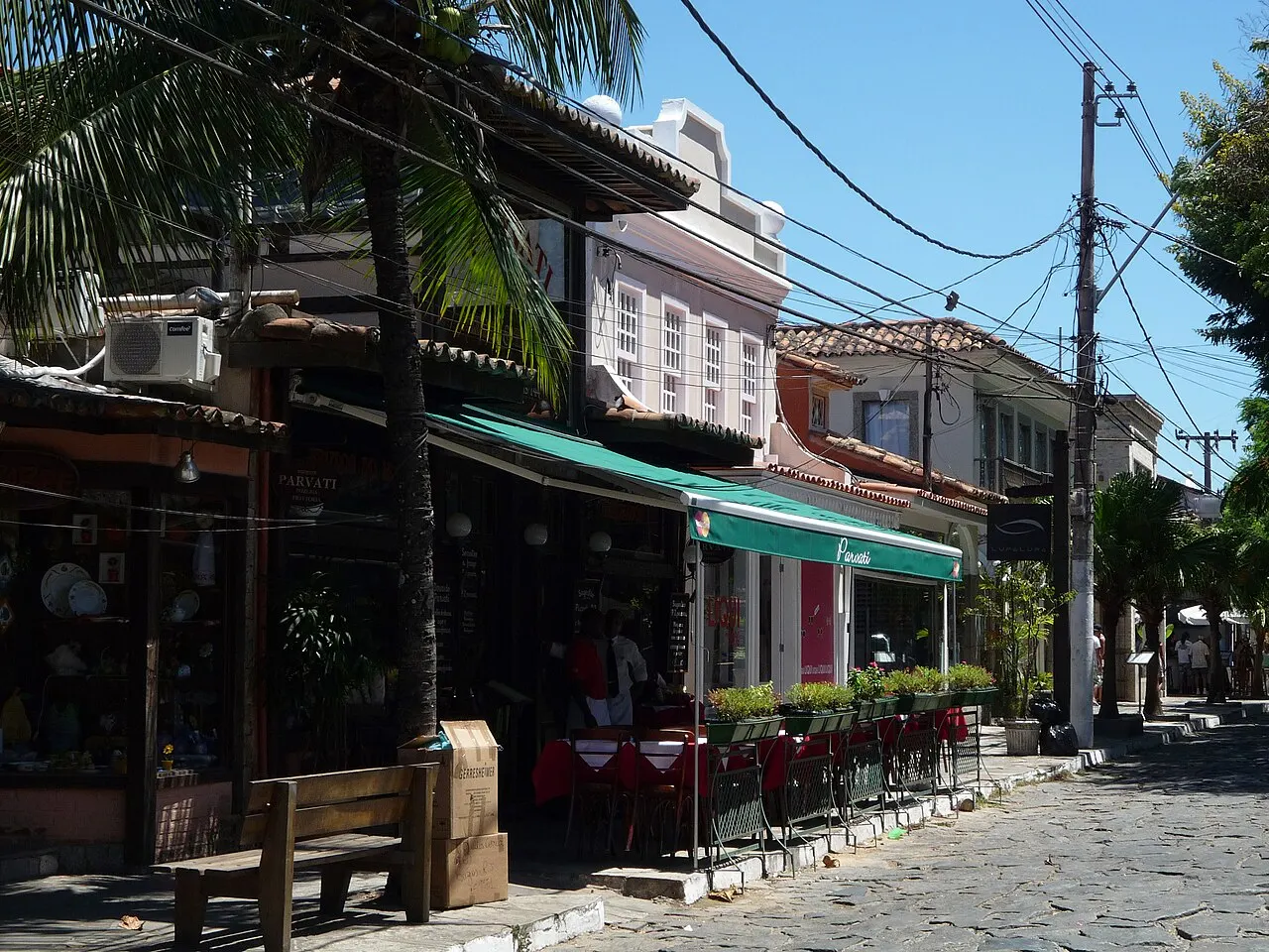 A Rua das Pedras, de calçamento de pedra, com mesas de bar e fachadas de lojas sob palmeiras.