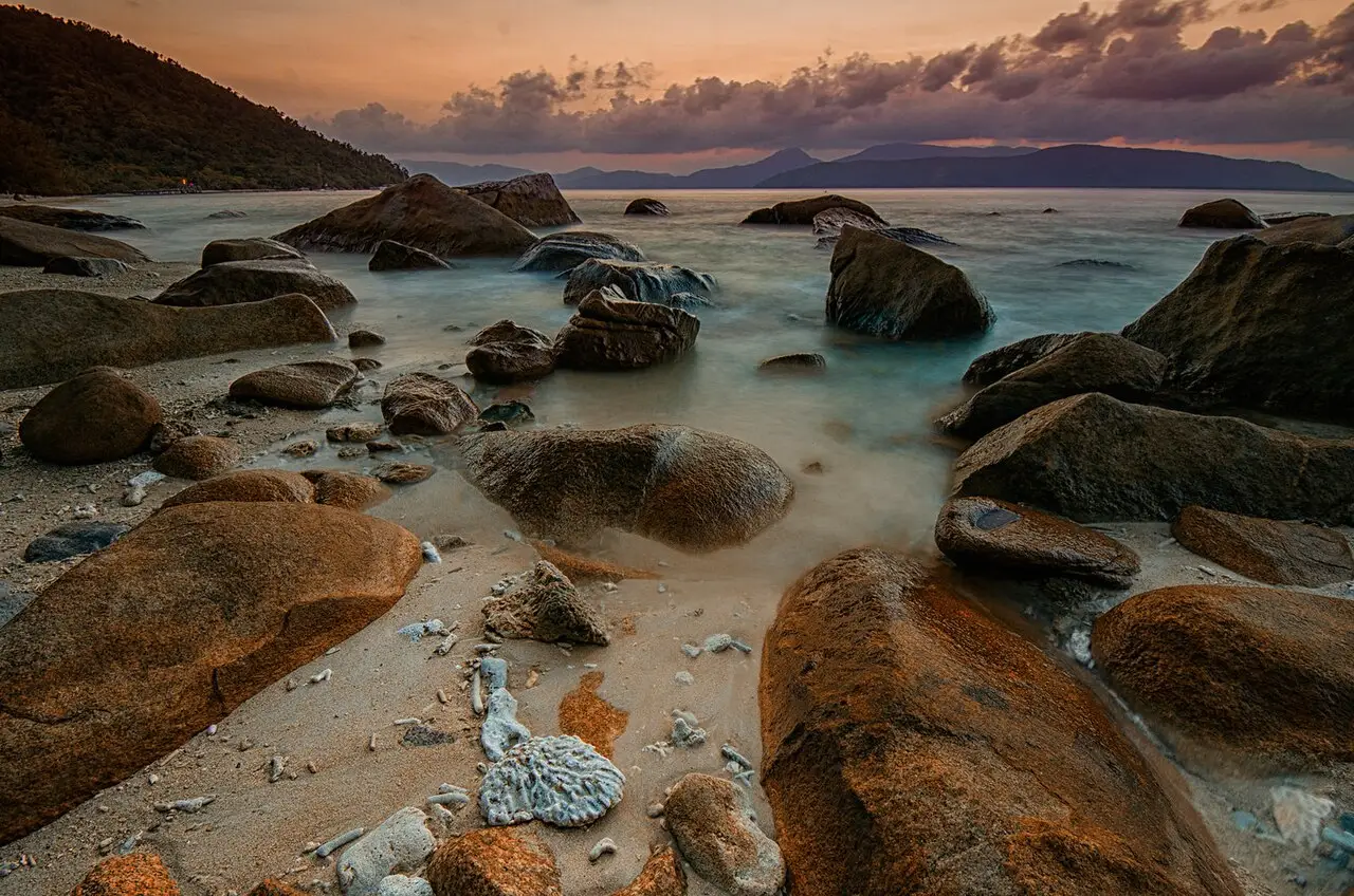 Costa da ilha Fitzroy, com pedras grandes na água rasa, um morro à esquerda e o mar ao fundo.