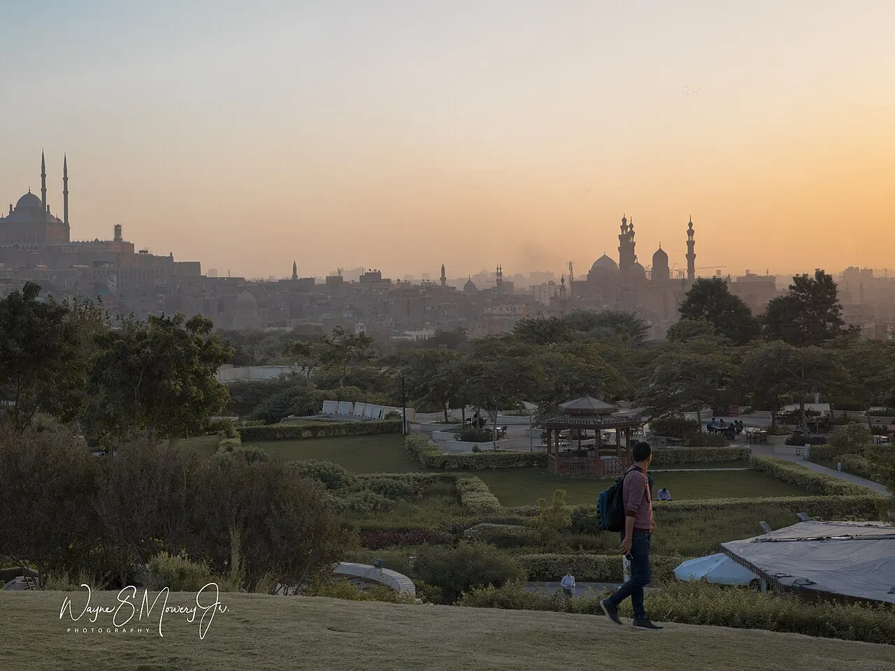 Os gramados e as sebes do Al-Azhar Park descem em planos, com uma pessoa no caminho e os minaretes da cidade sob um céu alaranjado.