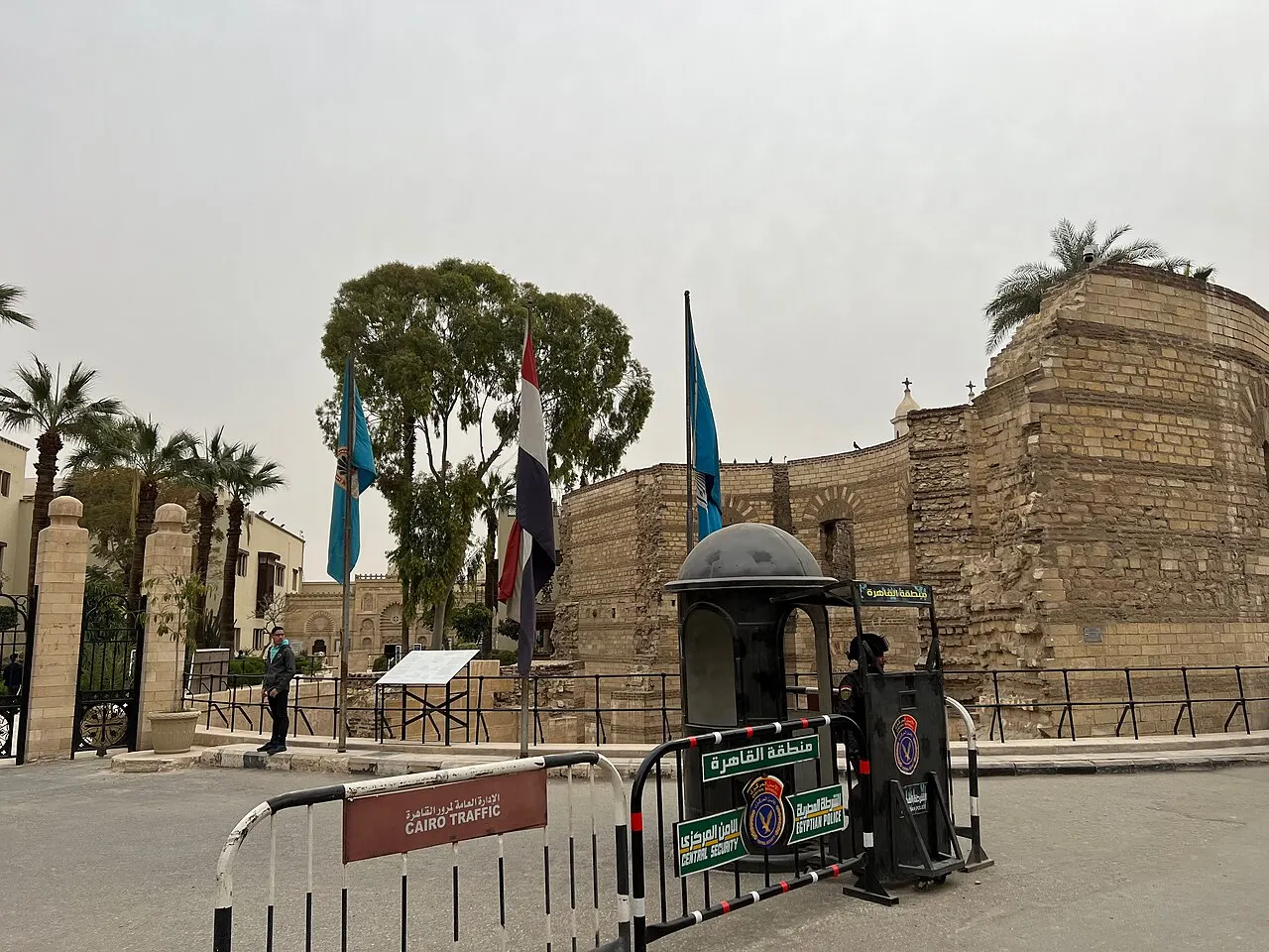 A torre redonda de pedra e a muralha do Cairo Copta ficam atrás de palmeiras, bandeiras e uma guarita.