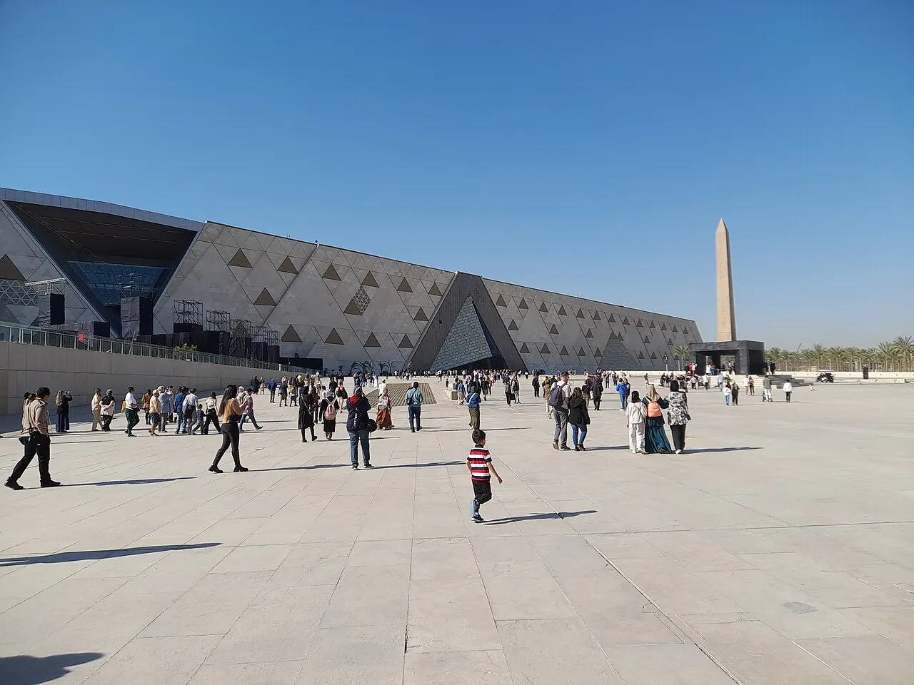 A fachada triangular de um grande museu, com painéis de pedra em padrão geométrico, um obelisco isolado na praça pavimentada em frente e visitantes caminhando pelo espaço aberto, sob céu azul.