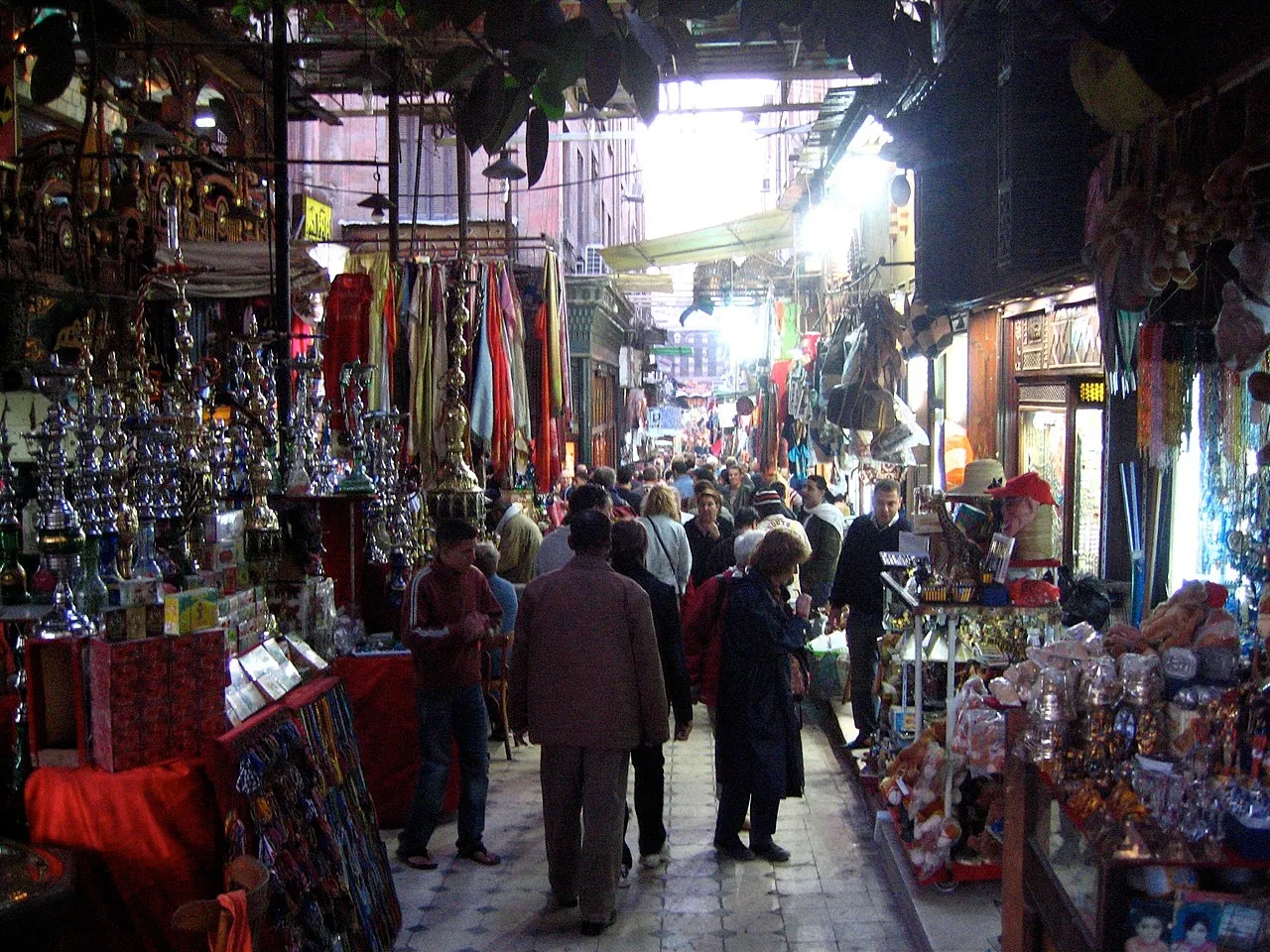 Um corredor coberto do Khan el-Khalili reúne lustres e metal à esquerda, tecidos à direita e gente caminhando no meio.