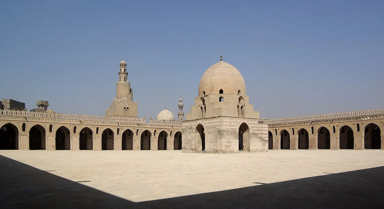Um grande pátio aberto cercado por arcadas de pedra em fileiras, com uma construção de cúpula sobre pilares ao centro e um minarete em espiral ao fundo, sob céu azul limpo.