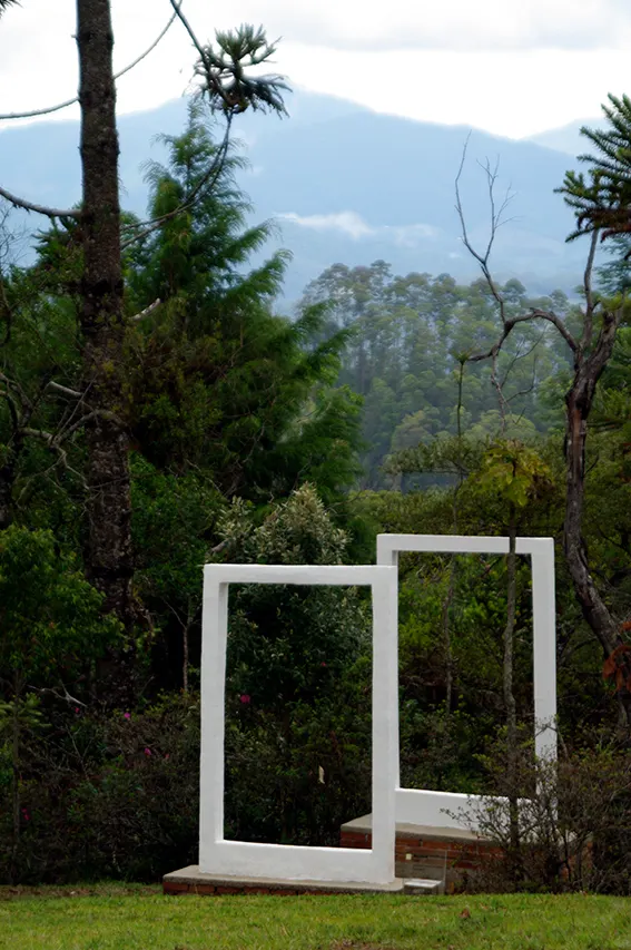 Uma escultura branca em forma de moldura sobre o gramado do Museu Felícia Leirner, com as árvores e a serra ao fundo.