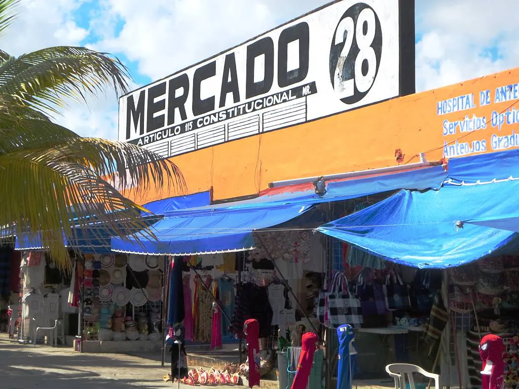 A fachada do Mercado 28, em Cancún, com o grande letreiro branco do mercado, toldos azuis e lojas de roupas e artesanato coloridos na calçada, com uma palmeira ao lado.