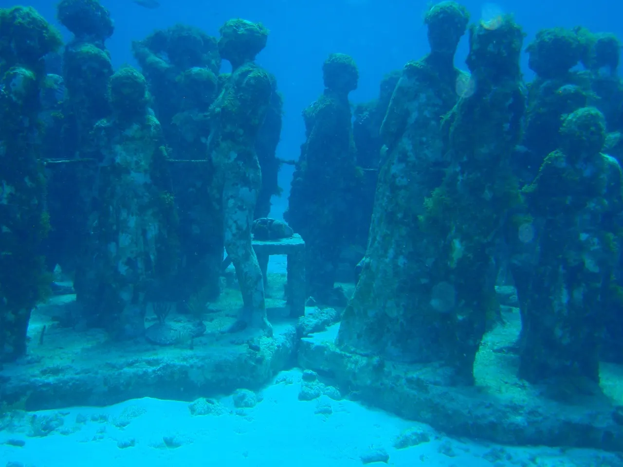 Dezenas de esculturas de figuras humanas em pé, cobertas de algas, no fundo de areia do mar, vistas embaixo d'água na luz azul do MUSA.