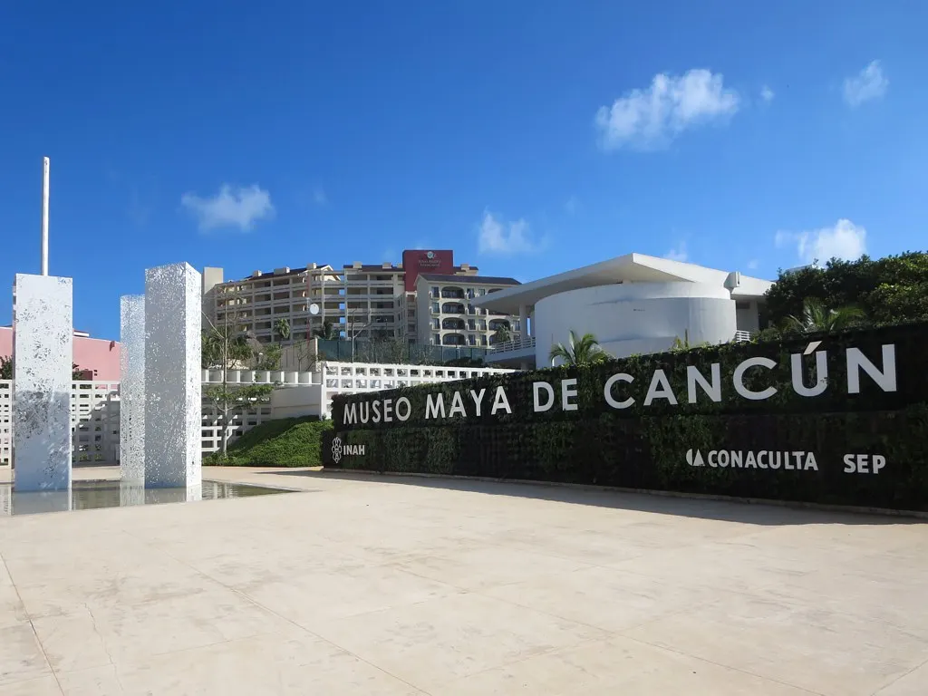 A entrada do Museo Maya de Cancún, com o nome do museu em letras brancas sobre um muro escuro, uma esplanada clara e o prédio branco do museu ao fundo, sob céu azul.