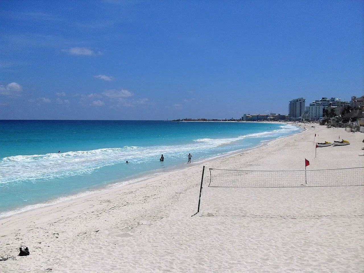 A Playa Delfines, em Cancún, com areia branca larga, mar turquesa com ondas, uma rede de vôlei, poucas pessoas na água e prédios de hotel no horizonte.