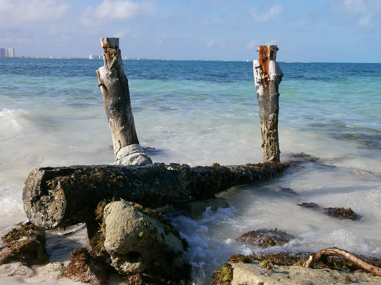 Dois troncos verticais e um tronco deitado entre pedras na beira da Playa Langosta, com a água rasa e clara da baía e a costa ao fundo.