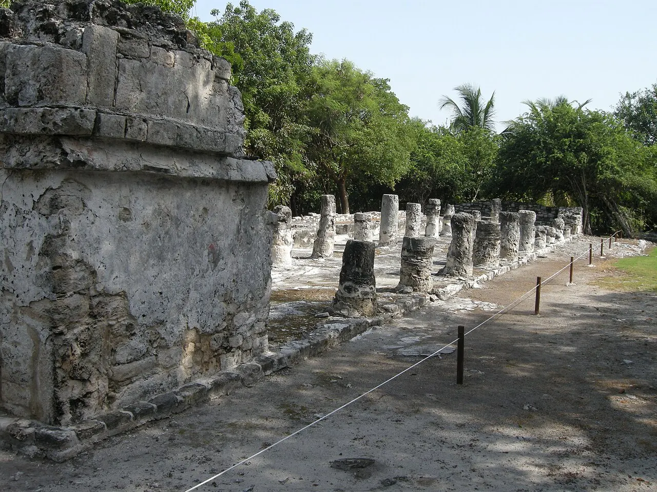 Uma fileira de colunas de pedra de uma construção maia em El Meco, ao lado da parede de um templo, com um caminho de cascalho, cordas de proteção e árvores ao fundo.