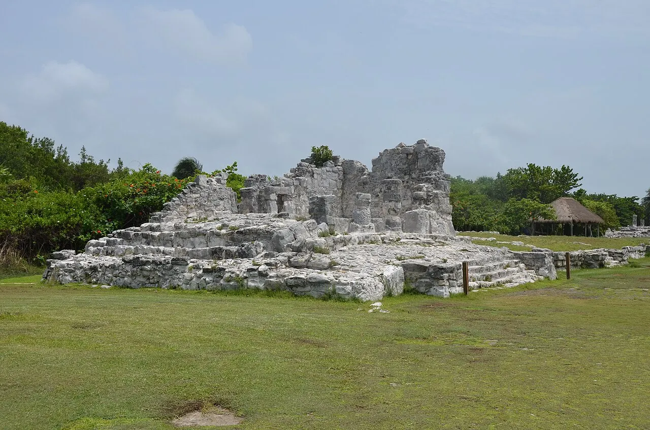 Uma estrutura maia de pedra calcária clara, em ruína, sobre uma plataforma em degraus, no sítio arqueológico El Rey, com gramado na frente e mata baixa atrás.