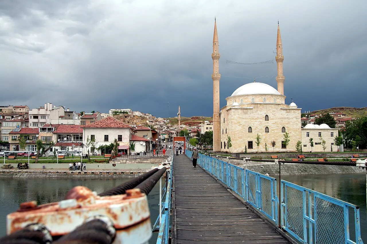 A ponte de pedestres com grade azul sobre o rio Kızılırmak, em Avanos, levando à cidade, com uma mesquita de dois minaretes e céu carregado.