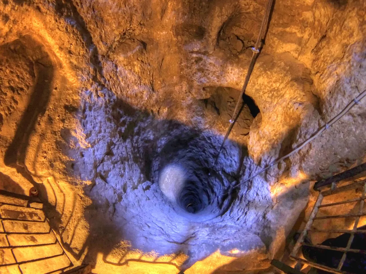 Um poço de ventilação da cidade subterrânea de Derinkuyu visto de cima, escavado na rocha e iluminado em tons de laranja e azul.