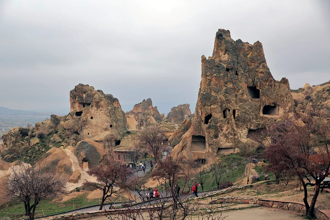 Formações de rocha clara do Museu ao Ar Livre de Göreme, com aberturas de mosteiros e igrejas escavadas, árvores sem folhas e visitantes na trilha.