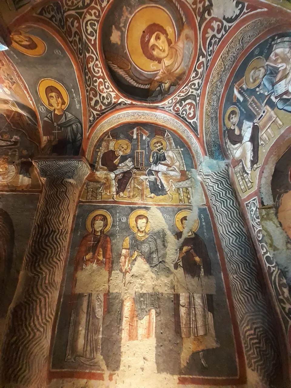 Afrescos no interior da Igreja Escura, em Göreme, com figuras de santos, auréolas douradas, arcos pintados e faixas ornamentais vermelhas, brancas e escuras.