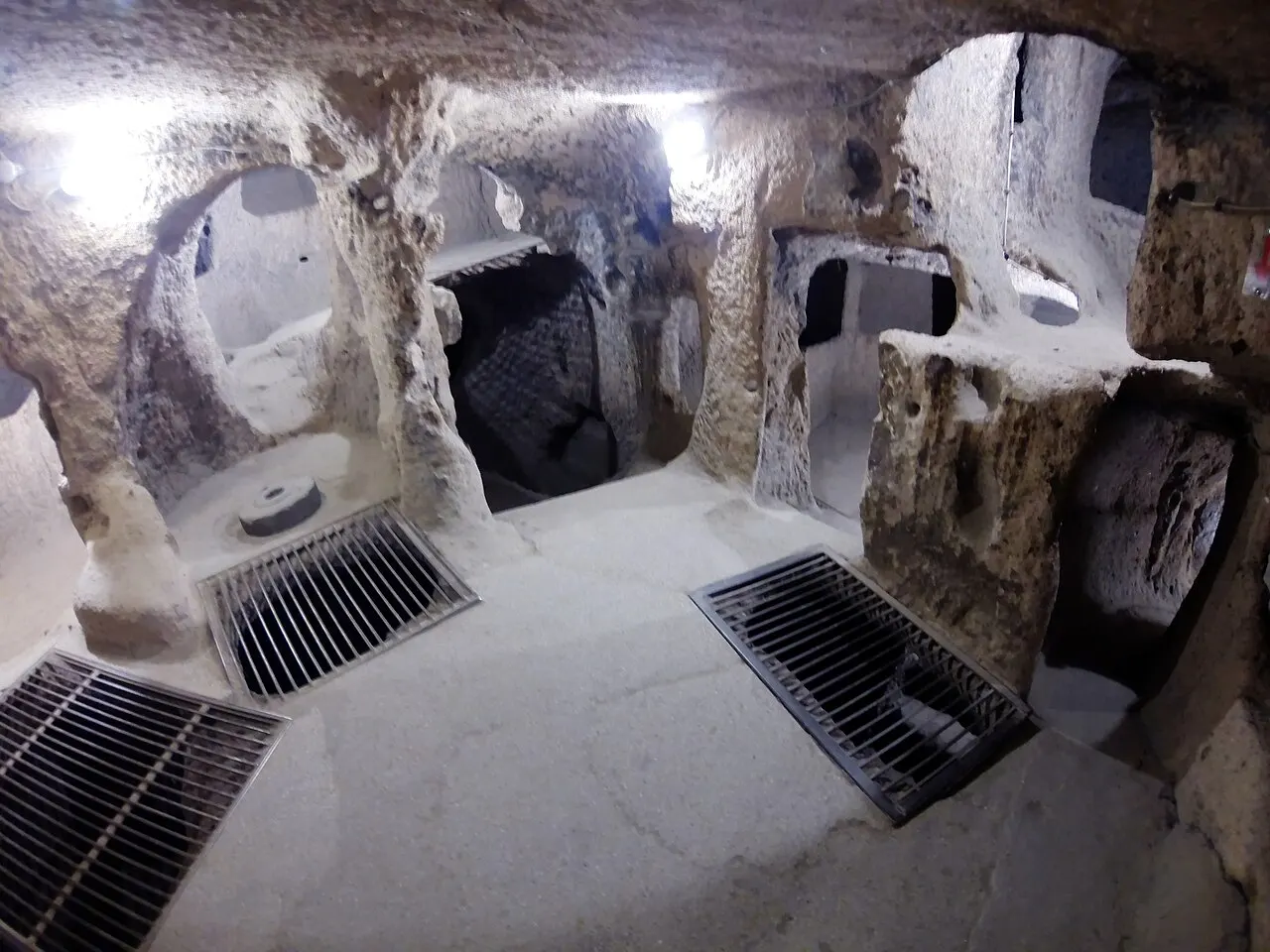 Uma sala da cidade subterrânea de Kaymaklı, com paredes de rocha clara, aberturas para outros cômodos e grades de proteção no chão.