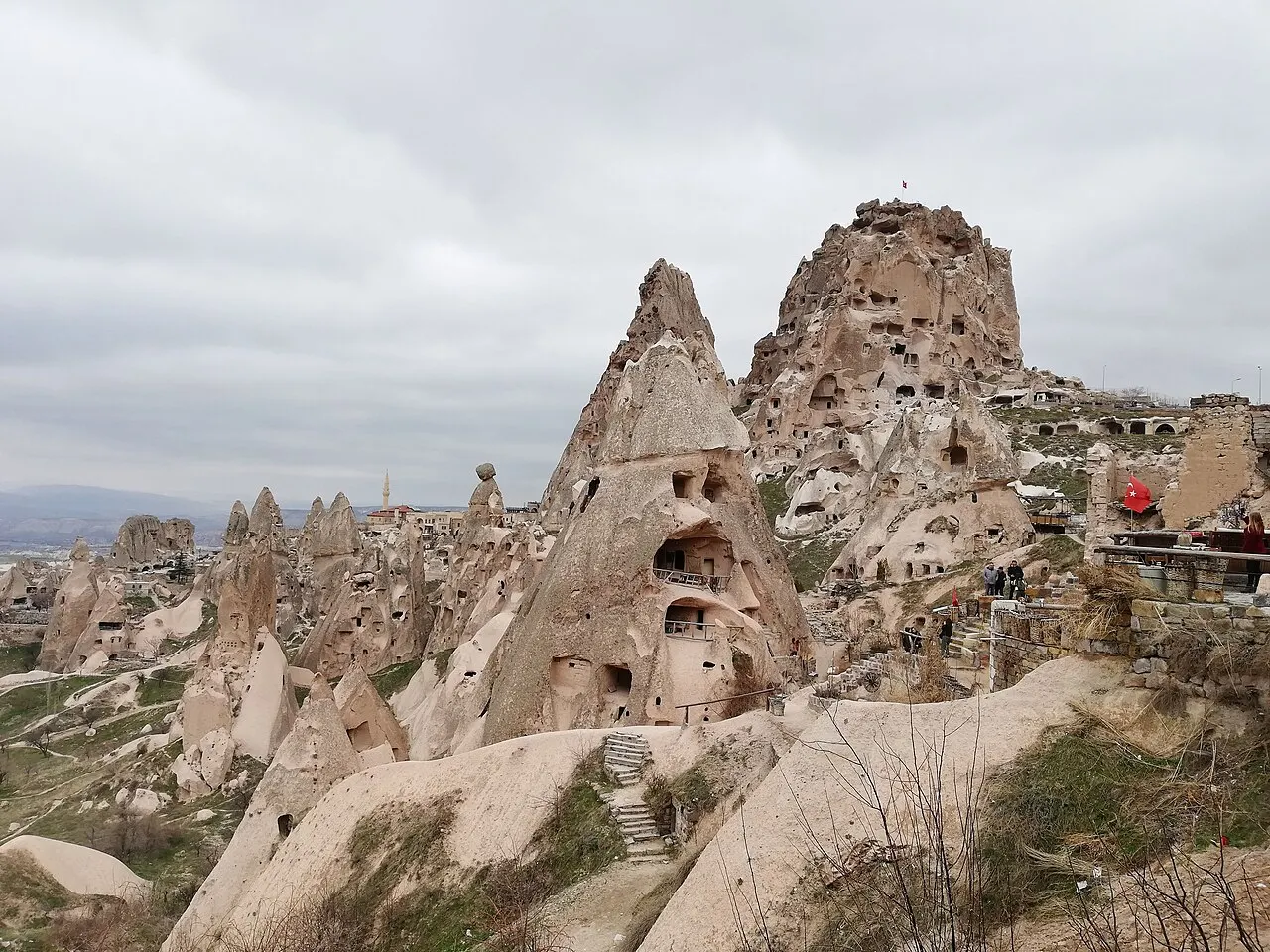 O rochedo do castelo de Uçhisar, um grande pico de pedra com cavidades escavadas, acima de chaminés de fada e casas, sob céu nublado.